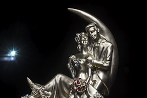 A metallic sculpture of a couple seated together, intricately designed with detailed features and decorative elements. The figures are positioned against a crescent moon backdrop, with elegantly sculpted attire adorned with small jewel-like details. The background is a solid black, enhancing the focus on the sculpture.