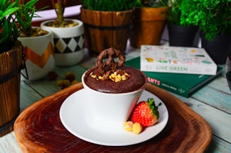 A chocolate dessert in a white ramekin on a wooden tray, garnished with chocolate pieces and chopped nuts. There is a fresh strawberry and a couple of hazelnuts beside the ramekin. Surrounding the dessert are potted plants and books, creating a casual and cozy atmosphere.