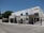 A modern, rectangular office building with a flat roof and large glass windows. The building is labeled 'Centro Empresarial' and features a gated entrance with metal fences. The surroundings include a wide street and some trees, indicating a suburban or business park location. The sky is clear and blue, suggesting a sunny day.