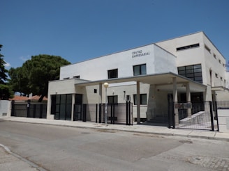 A modern, rectangular office building with a flat roof and large glass windows. The building is labeled 'Centro Empresarial' and features a gated entrance with metal fences. The surroundings include a wide street and some trees, indicating a suburban or business park location. The sky is clear and blue, suggesting a sunny day.