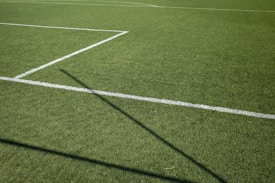 A section of a green grass sports field featuring intersecting white lines. The sunlight casts a shadow across the field, adding contrast to the vibrant grass surface.