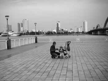 A parent is crouched down next to a small child in a stroller on a spacious, tiled waterfront promenade. The background features a river with several high-rise buildings and a large bridge. The scene is peaceful with no crowd presence and has a sense of stillness.