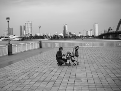 A parent is crouched down next to a small child in a stroller on a spacious, tiled waterfront promenade. The background features a river with several high-rise buildings and a large bridge. The scene is peaceful with no crowd presence and has a sense of stillness.