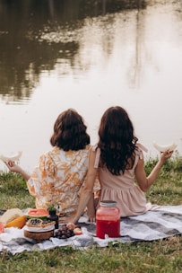 two women sitting on a blanket next to a body of water