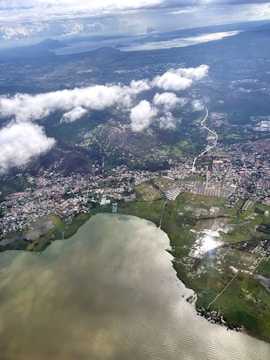An aerial view of a coastal region with a mix of urban and rural landscapes, featuring a body of water on the lower part of the image. Green fields and clusters of buildings are visible, along with winding roads connecting different areas. White clouds partially obscure sections of the land, while sunlight reflects on the water surface.