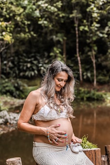 A pregnant woman is sitting outdoors, holding a small pair of baby shoes in one hand and her belly with the other. She is wearing a white lace crop top and a long skirt, accessorized with colorful beaded bracelets and a necklace. The background features lush greenery and a calm pond, creating a serene and natural setting.