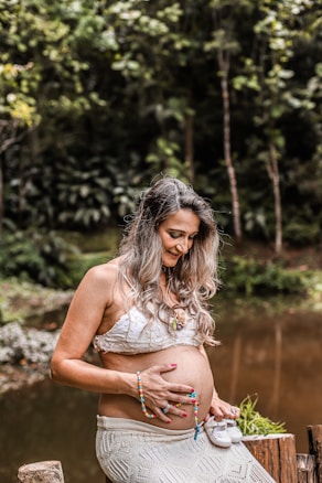 A pregnant woman is sitting outdoors, holding a small pair of baby shoes in one hand and her belly with the other. She is wearing a white lace crop top and a long skirt, accessorized with colorful beaded bracelets and a necklace. The background features lush greenery and a calm pond, creating a serene and natural setting.