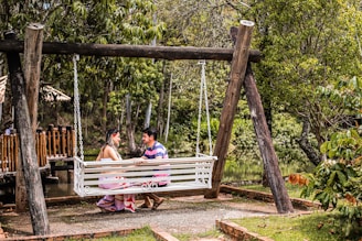 A couple is seated on a white, wooden swing suspended between two large wooden posts in a lush garden setting. They are facing each other, engaged in an intimate conversation. Surrounding them are green trees and other vegetation, and a wooden fence and pond are visible in the background. The scene is peaceful and natural.