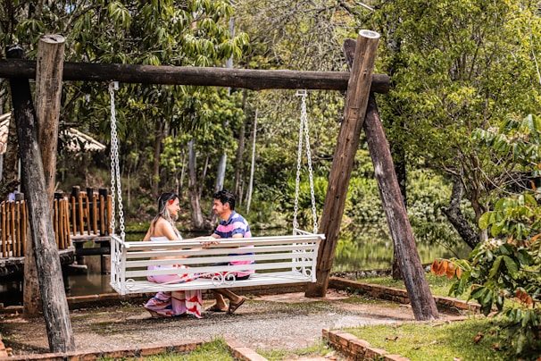 A couple is seated on a white, wooden swing suspended between two large wooden posts in a lush garden setting. They are facing each other, engaged in an intimate conversation. Surrounding them are green trees and other vegetation, and a wooden fence and pond are visible in the background. The scene is peaceful and natural.