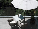 A patio or balcony area featuring a simple outdoor setup with a square table and two wicker chairs. A large white umbrella provides shade. There is greenery visible in the background behind a glass railing, creating a tranquil outdoor atmosphere.