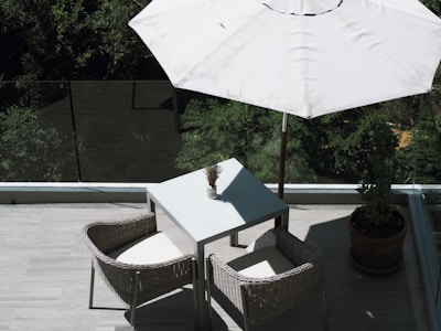 A patio or balcony area featuring a simple outdoor setup with a square table and two wicker chairs. A large white umbrella provides shade. There is greenery visible in the background behind a glass railing, creating a tranquil outdoor atmosphere.