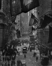 A bustling city street filled with people celebrating, with confetti raining down in a lively commemoration. Numerous American flags are prominently displayed, hanging from the buildings that line the street. The scene is black and white, capturing an historic parade or event, likely in a major city like New York.
