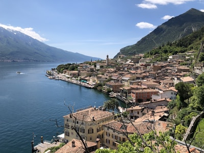 A picturesque lakeside town nestled between lush green hills and mountains. The town features a collection of closely packed, colorful buildings with terracotta roofs. The serene lake reflects the blue sky, with a small boat visible in the water. Vegetation and trees are abundant, contributing to the tranquil and scenic atmosphere.