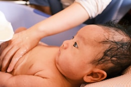 A baby with wet hair is lying in a plastic bath. An adult hand gently touches the baby's chest, and water is being poured over the baby from a small white container. The scene is intimate and shows a bathing session.