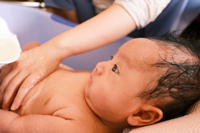 A baby with wet hair is lying in a plastic bath. An adult hand gently touches the baby's chest, and water is being poured over the baby from a small white container. The scene is intimate and shows a bathing session.