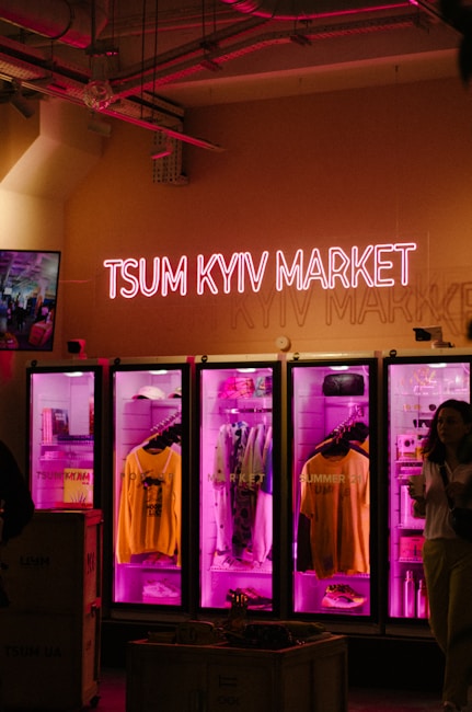 A neon sign reading 'TSUM Kyiv Market' is mounted above a row of three brightly lit display cases. Each display case is illuminated with vibrant pink lighting, showcasing various clothing items. A person on the right holds a cup, partially visible in the image's shadowy area, adding a dynamic, lived-in feel to the scene.