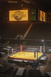 An indoor arena with rows of stadium seating and a central boxing ring under bright lights. Above the ring, a large electronic sign displays the name 'Butte Civic Center'. The arena is mostly empty, with only a few people seen near the ring and some scattered chairs and tables.