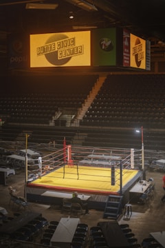 An indoor arena with rows of stadium seating and a central boxing ring under bright lights. Above the ring, a large electronic sign displays the name 'Butte Civic Center'. The arena is mostly empty, with only a few people seen near the ring and some scattered chairs and tables.