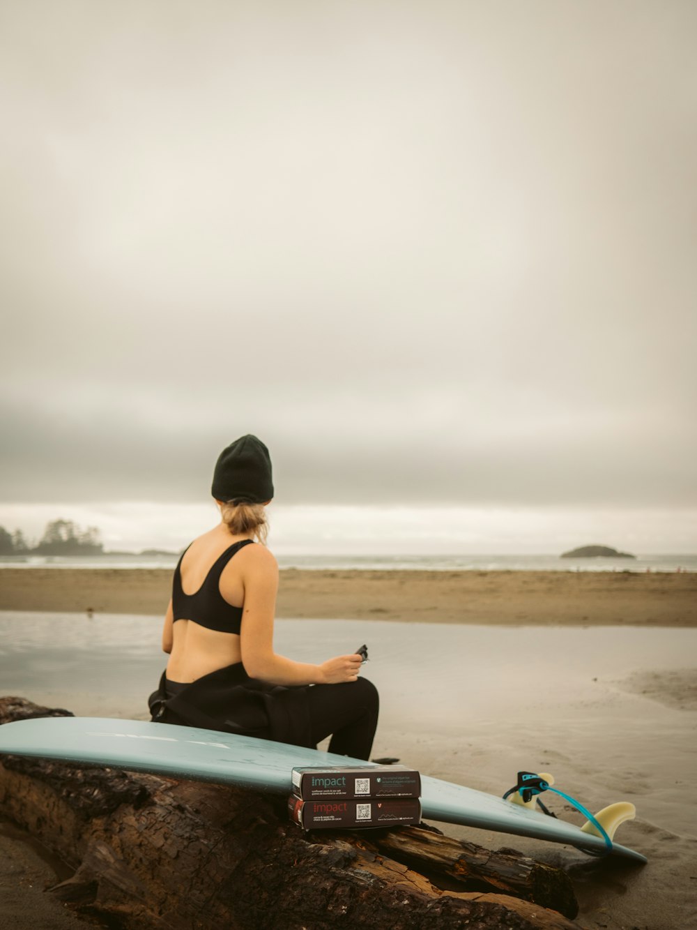 uma mulher sentada em um tronco ao lado de uma prancha de surf