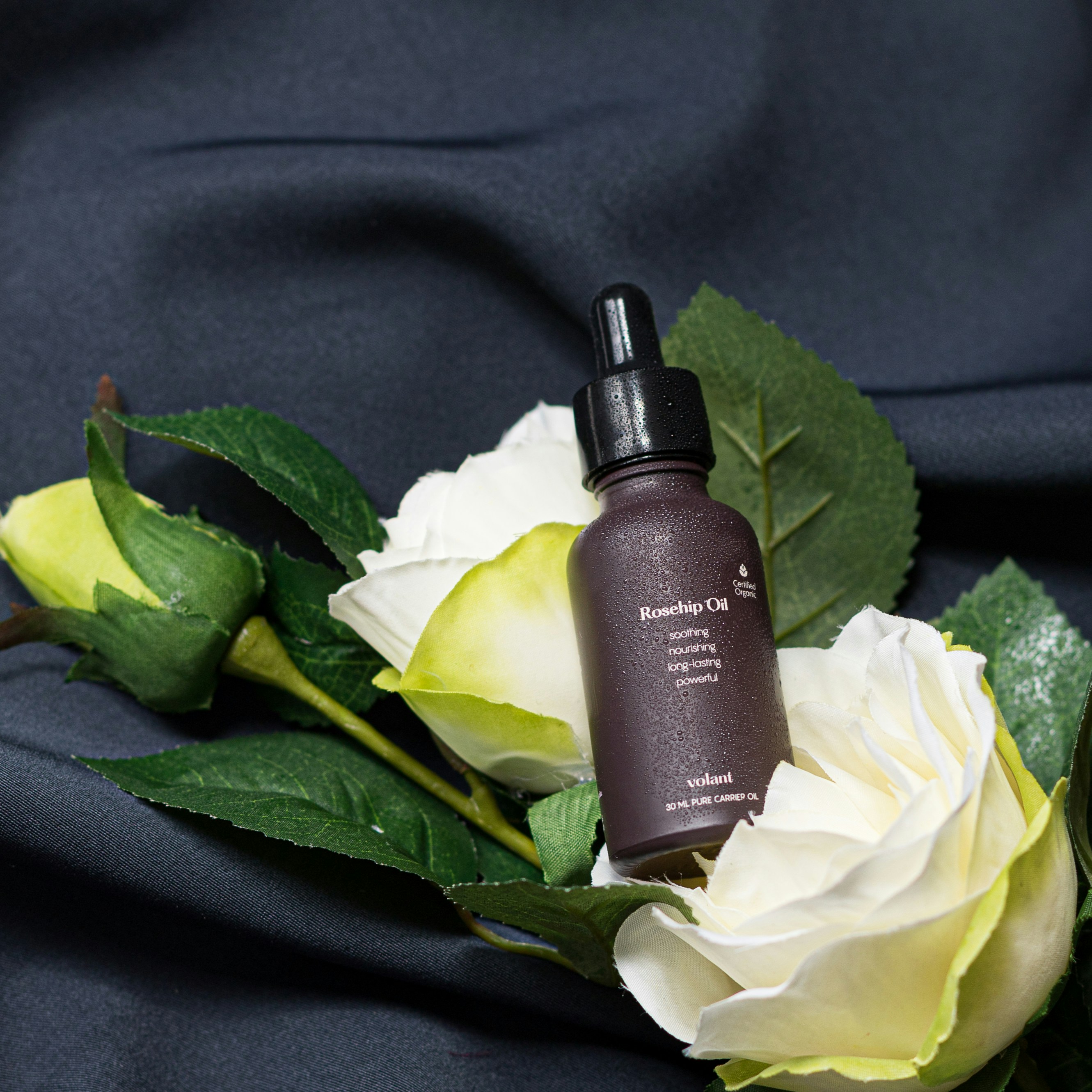 Amber glass bottle of rosehip oil surrounded by dried rose petals on cream linen fabric