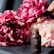 A dropper is dispensing oil into a small dark brown bottle labeled 'Jojoba Oil,' surrounded by lush pink and white peonies in the background. The scene suggests a focus on natural beauty and skincare.