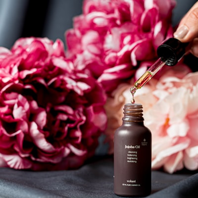 A dropper is dispensing oil into a small dark brown bottle labeled 'Jojoba Oil,' surrounded by lush pink and white peonies in the background. The scene suggests a focus on natural beauty and skincare.