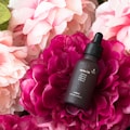 A dark brown dropper bottle labeled 'Jojoba Oil' is surrounded by vibrant pink and white peonies. The bottle has a textured surface, and water droplets are visible on its exterior, creating a fresh and appealing look.