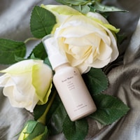 A small bottle labeled 'Rose Water' is surrounded by creamy white roses with lush green leaves. The setting includes a textured gray fabric that adds elegance to the arrangement.