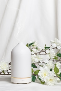 A white ceramic diffuser is placed on a white surface, surrounded by delicate white flowers and green leaves. A soft mist is emerging from the top of the diffuser, creating a calm and serene atmosphere. The background is a light fabric that complements the overall clean and minimalist aesthetic.