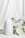 A white ceramic diffuser is placed on a white surface, surrounded by delicate white flowers and green leaves. A soft mist is emerging from the top of the diffuser, creating a calm and serene atmosphere. The background is a light fabric that complements the overall clean and minimalist aesthetic.