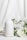 A white ceramic diffuser is placed on a white surface, surrounded by delicate white flowers and green leaves. A soft mist is emerging from the top of the diffuser, creating a calm and serene atmosphere. The background is a light fabric that complements the overall clean and minimalist aesthetic.