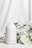 A white ceramic diffuser is placed on a white surface, surrounded by delicate white flowers and green leaves. A soft mist is emerging from the top of the diffuser, creating a calm and serene atmosphere. The background is a light fabric that complements the overall clean and minimalist aesthetic.