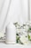 A white ceramic diffuser is placed on a white surface, surrounded by delicate white flowers and green leaves. A soft mist is emerging from the top of the diffuser, creating a calm and serene atmosphere. The background is a light fabric that complements the overall clean and minimalist aesthetic.