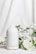 A white ceramic diffuser is placed on a white surface, surrounded by delicate white flowers and green leaves. A soft mist is emerging from the top of the diffuser, creating a calm and serene atmosphere. The background is a light fabric that complements the overall clean and minimalist aesthetic.