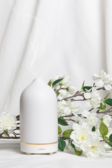 A white ceramic diffuser is placed on a white surface, surrounded by delicate white flowers and green leaves. A soft mist is emerging from the top of the diffuser, creating a calm and serene atmosphere. The background is a light fabric that complements the overall clean and minimalist aesthetic.
