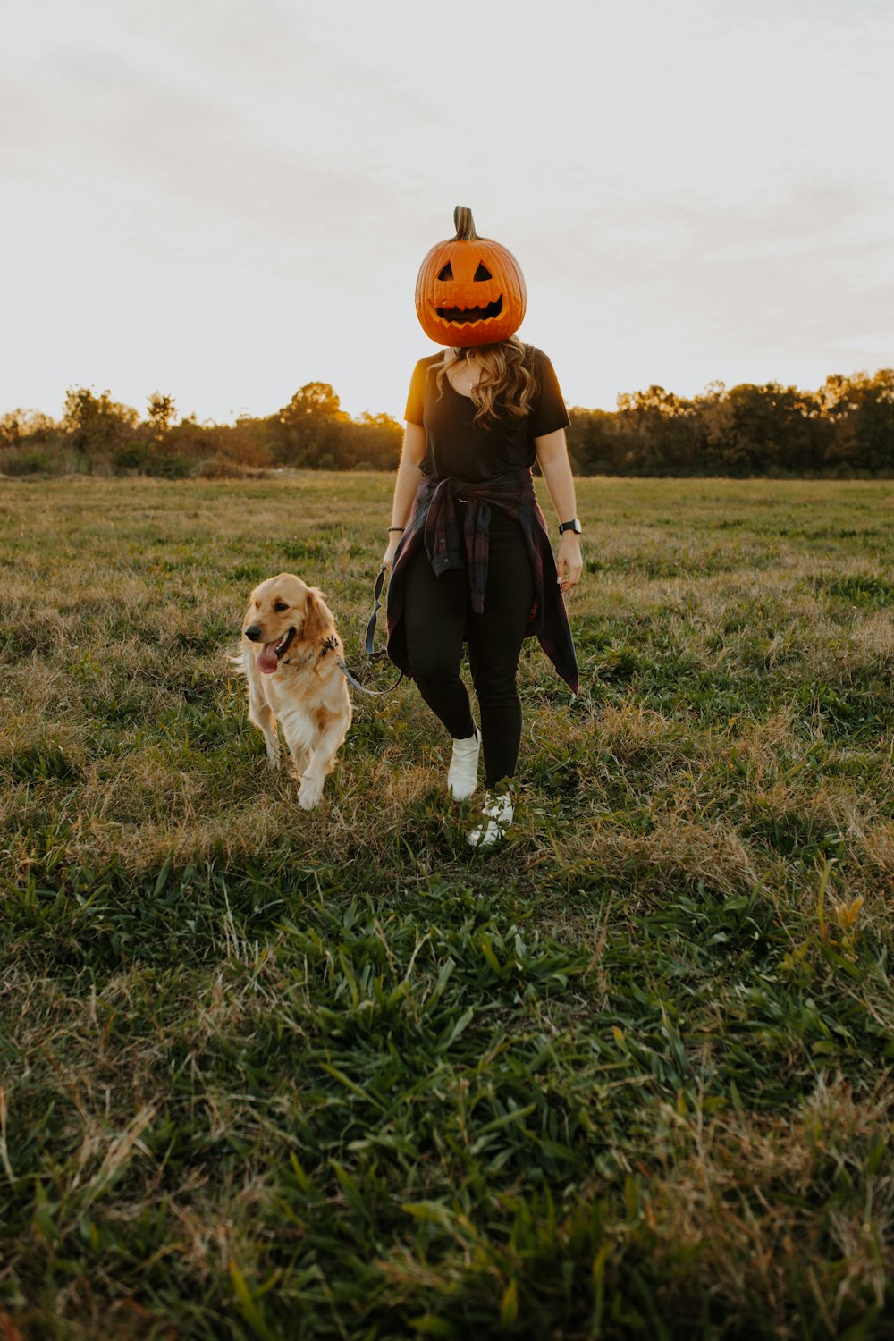 a woman walking a dog in a field with a pumpkin on her head