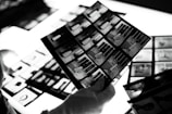 A hand wearing a white glove is holding negatives or photo contact sheets with multiple black and white images. The background includes more sheets on a bright lightbox, which enhances the details.