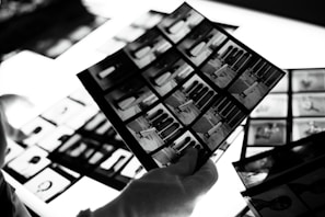 A hand wearing a white glove is holding negatives or photo contact sheets with multiple black and white images. The background includes more sheets on a bright lightbox, which enhances the details.