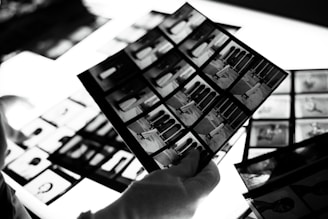 A hand wearing a white glove is holding negatives or photo contact sheets with multiple black and white images. The background includes more sheets on a bright lightbox, which enhances the details.