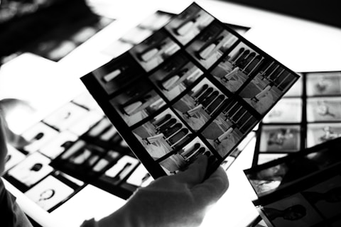 A hand wearing a white glove is holding negatives or photo contact sheets with multiple black and white images. The background includes more sheets on a bright lightbox, which enhances the details.