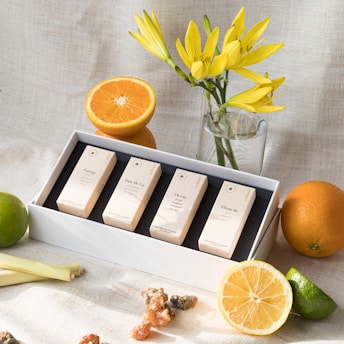 A display of four boxed fragrances set in a white tray, surrounded by fresh citrus fruits including oranges, lemons, and limes. Yellow flowers in a glass vase add a touch of freshness, and the background is a light, textured fabric.