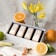 A display of four boxed fragrances set in a white tray, surrounded by fresh citrus fruits including oranges, lemons, and limes. Yellow flowers in a glass vase add a touch of freshness, and the background is a light, textured fabric.