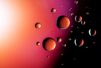 Multiple droplets of liquid appear to be suspended against a gradient background transitioning from light orange to dark purple. The droplets are reflective and exhibit a glossy finish, each displaying a spectrum of colors from the surrounding ambient light.