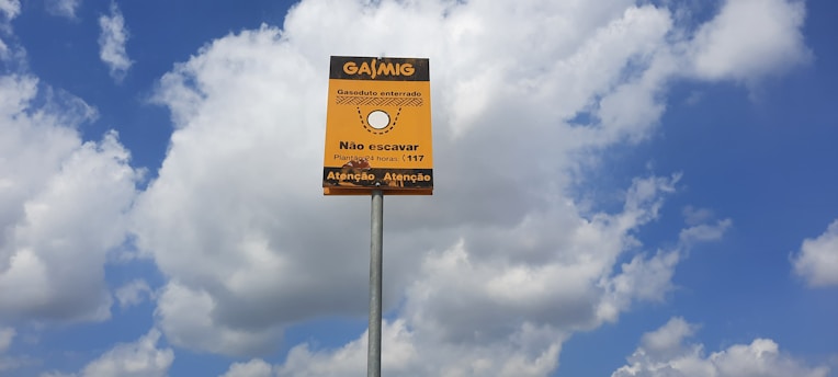 A yellow sign with black text in Portuguese stands against a backdrop of a blue sky with scattered white clouds. The sign appears to be a warning about an underground gas pipeline, advising not to dig. It is mounted on a tall metal pole.