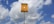 A yellow sign with black text in Portuguese stands against a backdrop of a blue sky with scattered white clouds. The sign appears to be a warning about an underground gas pipeline, advising not to dig. It is mounted on a tall metal pole.
