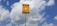 A yellow sign with black text in Portuguese stands against a backdrop of a blue sky with scattered white clouds. The sign appears to be a warning about an underground gas pipeline, advising not to dig. It is mounted on a tall metal pole.