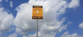 A yellow sign with black text in Portuguese stands against a backdrop of a blue sky with scattered white clouds. The sign appears to be a warning about an underground gas pipeline, advising not to dig. It is mounted on a tall metal pole.