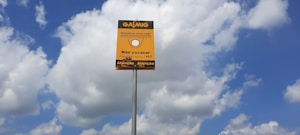 A yellow sign with black text in Portuguese stands against a backdrop of a blue sky with scattered white clouds. The sign appears to be a warning about an underground gas pipeline, advising not to dig. It is mounted on a tall metal pole.