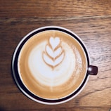A cup of coffee with intricate latte art in the shape of a heart and leaf pattern, set on a wooden table. The coffee is in a brown cup with a saucer that matches.