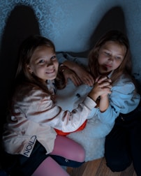 Two young girls are sitting on a floor with their hands clasped together in a gesture of friendship or affection. They are leaning against a fluffy white pillow, with one of them smiling directly at the viewer. The room is dimly lit, creating soft shadows against the patterned wallpaper.